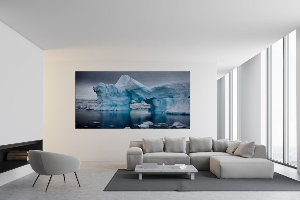 Ein Fotokunstwerk hängt an der Wand in einem Wohnzimmer. Auf dem Foto ist ein Eisberg zu sehen. Er ist umgeben von Wasser und winzigen Eisschollen. Das Bild ist in der Antarktis entstanden.